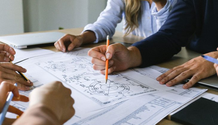 Hands discussing engineering plans on a table with detailed diagrams and notes.